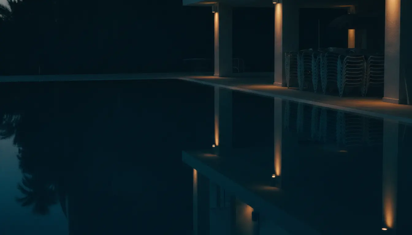 A resort pool terrace after dark: still water reflecting lit colonnades and stacked loungers under the shelter.